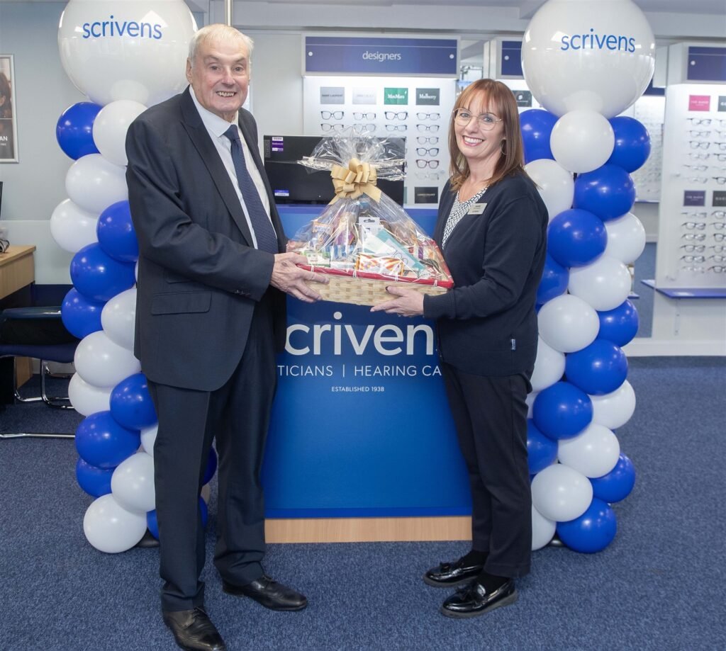 Ken Taylor and branch manager Jeanette Green holding a luxury hamper in front of a blue and white Scrivens balloon arch